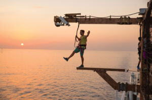 A daring man stands on one legs on a narrow beam that protrudes for the deck of a cruise ship.